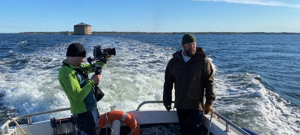 En kameraman och skådespelare i båt på havet. I bakgrunden syns ett kastell.